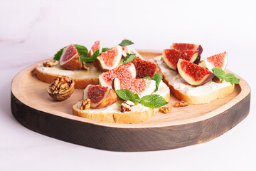 Tasty homemade sandwiches with sweet figs, cream cheese, walnuts. honey and mint leaves on the wooden serving plate on light marble background close up.