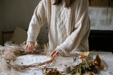 Woman making stylish autumn wreath. Hands with dried grass, herbs, leaves, ribbon and scissors on rustic table. Details of fall decor and arrangement in home