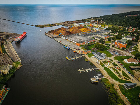 An aerial view of Salacgriva ship port, showcasing the marina with docks for small boats, a lighthouse, and surrounding greenery. The port area features well maintained pathways, parked vehicles