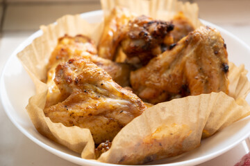 Homemade meal cooked in air fryer grill. Freshly fried chicken wings are placed on paper in white plate with the light shining through the window falling on the chicken. Selective focus