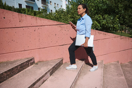 Confident businesswoman carrying a laptop walks up outdoor stairs while checking her phone