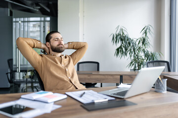 Fototapeta premium Businessman taking relaxing break at desk with laptop, hands behind head, eyes closed, and documents nearby in modern office. Depicts stress-free work environment, productivity, and comfort