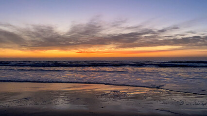 Fototapeta premium Orange and pink skies as sun goes down overlooking sandy beach while ocean tides create waves