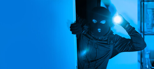 A person dressed in dark clothing and a ski mask peers into a dimly lit room while holding a...