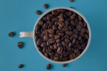 A coffee cup filled with roasted coffee beans with a blue background