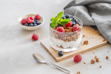 Greek yogurt with granola, raspberries and blueberries in a glass on a wooden board on a light background with fresh berries and  morning sunlight close up.