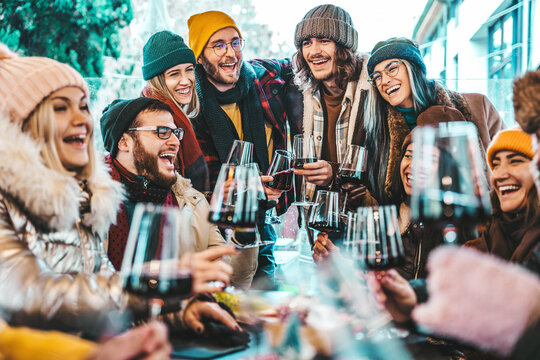 Happy multiracial friends toasting red wine at restaurant terrace - Group of young people wearing winter clothes having fun at outdoors winebar table - Dining life style and friendship concept