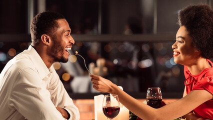 Joyful Black Couple Of Lovers Feeding Each Other During Romantic Date In Fancy Restaurant. Valentine's Romance And Relationship