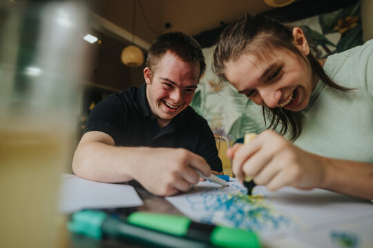Two young friends, a male with Down syndrome and a girl in a wheelchair, joyfully engage in drawing and painting together, expressing creativity and friendship.