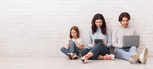 Family Gadgets. Happy parents and their little daughter holding different electronic devices in hands while sitting on floor near white brick wall
