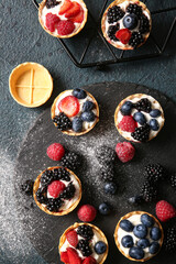 Board of tasty tartlets with whipped cream and berries on dark background