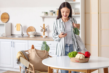 Young Asian woman with eco bags full of fresh food and mobile phone in kitchen