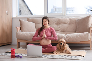Sporty young pregnant woman with laptop and cocker spaniel meditating at home
