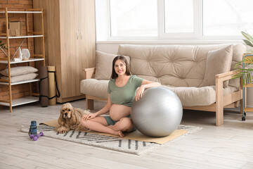 Beautiful sporty young pregnant woman with cocker spaniel and fitball practicing yoga at home