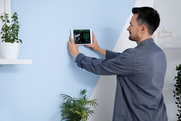 Young man with smart home security system control panel in bedroom, back view