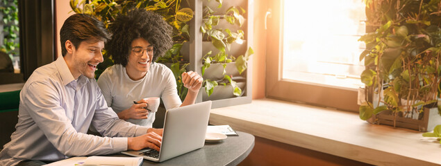 Two multiethnic guys colleagues collaborate while looking at a laptop in a sunlit cafe adorned with greenery, copy space