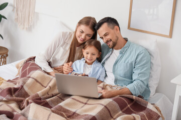 Little girl and her parents with warm plaid using laptop in bedroom