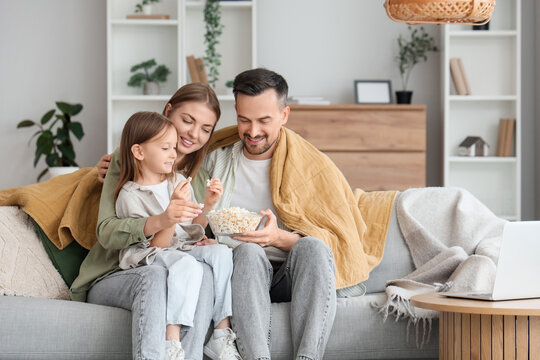 Little girl and her parents with warm plaid eating popcorn on sofa at home