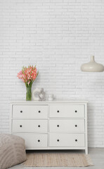 Vase with gladiolus flowers on commode near white brick wall in room interior