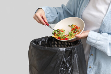 Young woman throwing organic food into trash bin on light background, closeup. Waste recycling concept