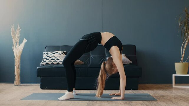 A woman performs a backbend yoga pose on a mat in her cozy living room. The space is bright, featuring a comfortable sofa and plants, creating a peaceful atmosphere for practice.