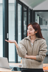 A young woman in a casual sweater stands in a modern office, gesturing with her hand, with a laptop and office supplies on the desk.