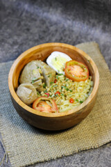 Meatball noodles are served in a round wooden bowl placed on a piece of burlap. top with chopped spring onions, sliced ​​red chilies and two slices of tomato.