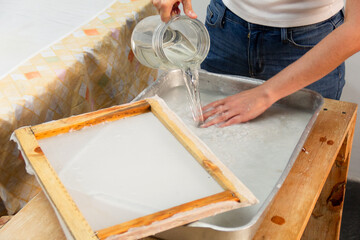 Close-up of person pouring water into a tray soaking paper and using deckle in the process of recycling paper in a workshop. Eco friendly and zero waste concept.