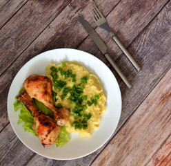A plate with two baked chicken legs and mashed potatoes on a wooden table with cutlery.