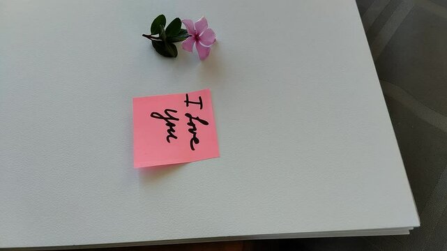 Una toma de cerca de una persona pegando con las manos una nota con la palabra te amo al lado de una flor rosada en una hoja de color blanco