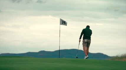 Male golfer reading putting green, reading putt near pin with club in hand on Ireland links golf course on beautiful sunny day, walking in slow motion