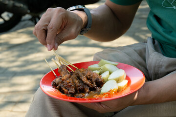 Sate Ayam Madura (Chicken Satay), an Indonesian traditional food that served alongside peanut sauce, soy sauce, and traditional rice cake (lontong). Sold on a street food stall, on melamine plate