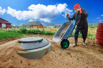Installing wastewater plumbing for country house, worker uses wheelbarrow to transport sand.