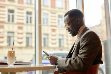 African American businessman using smartphone while sitting by window in modern cafe, drinking iced coffee. Sunlight reflecting on glass creating warm ambiance