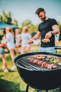 Un hombre cocina brochetas en una barbacoa mientras disfruta del verano al aire libre. Al fondo, otras personas crean una atm&oacute;sfera de picnic.