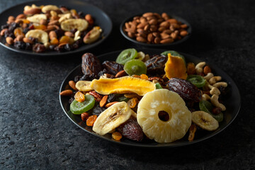 Dried tropical fruits, nuts, and raisins in black plate.