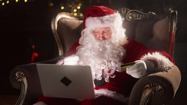 Online shopping and internet fraud, Santa Claus giving card data to scammers. Portrait of old man in costume of Santa sitting on armchair with laptop and credit card at cozy home, cyber security