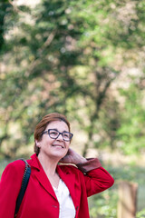 A mature woman with glasses smiles warmly while wearing a red blazer. The background is blurred, with lush green foliage and sunlight filtering through.