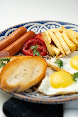 Breakfast plate with fried eggs, sausage, tomatoes, and fries served on a decorative blue plate