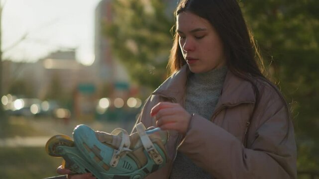 A young woman wearing a peach jacket and a gray sweater is seen adjusting the straps of her light blue rollerblades while standing in a park. The blurred background includes green trees