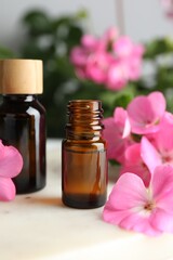 Bottle of geranium essential oil and beautiful flowers on white table, closeup