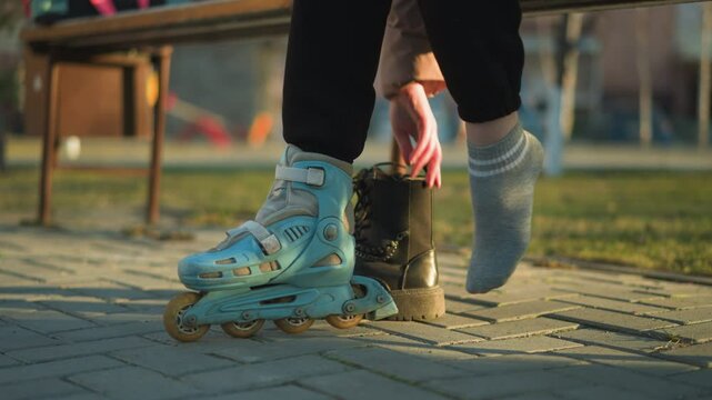 A person sitting in a park, with one leg in a rollerblade, pulling off a black boot from the other leg and preparing to wear the second rollerblade