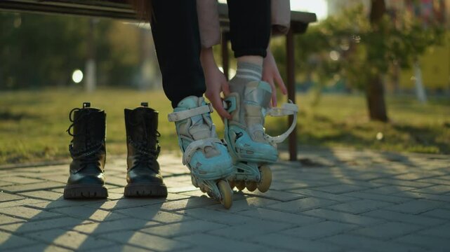 A person wearing a peach jacket and black trousers sits on a park bench, reaching for a rollerblade placed on the ground, preparing to put it on