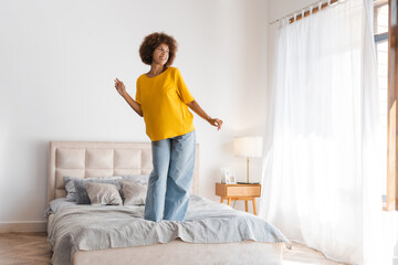 Smiling young pretty woman dancing standing on the bed in bedroom, looking out the window