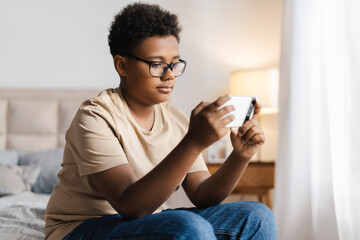 African American little boy in glasses looking at mobile phone playing game in bedroom