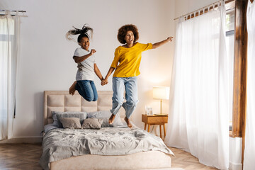 Mixed-race mother and daughter holding hands jumping on bed in bedroom