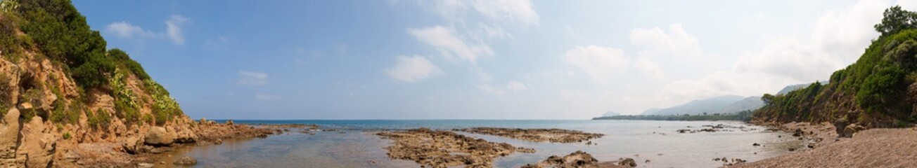 A wide panoramic view of a rocky coastal shoreline, featuring cliffs covered in greenery, cacti, and a calm sea under a bright sky. A serene and expansive coastal landscape
