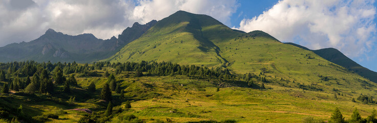 Obraz premium A wide panoramic view of rolling green hills leading up to towering mountain peaks under a partly cloudy sky, capturing the serene and expansive beauty of the alpine landscape