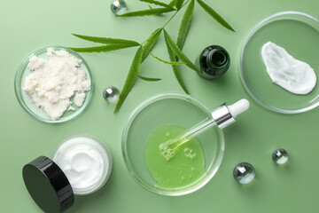 Petri dishes with cosmetic products, pipette and leaves on green background, flat lay
