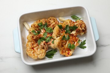 Tasty cauliflower steaks and parsley in baking dish on white marble table, top view